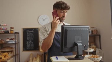 Fırında çalışan genç adam, arka planda hamur işleri olan tezgahın arkasındaki bilgisayarı kullanırken telefonla konuşuyor.