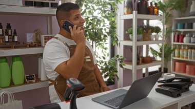 Akıllı telefonlu genç adam, önünde bir dizüstü bilgisayar ve raflarla iyi aydınlatılmış bir dekor dükkanının tezgahında konuşuyor.