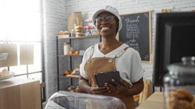 Bir pastanede önlük ve şapka takmış, elinde tablet, arkasında ekmek ve hamur işleri raflarıyla gülümseyen Afrikalı bir kadın..