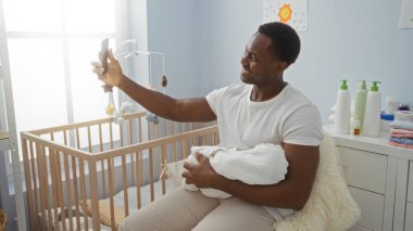 Genç adam, rahat bir yatak odasında selfie çekerken mutlu bir şekilde beşiği kucaklıyor. Huzurlu bir evde, sevgi dolu bir babalık anını yakalıyor..