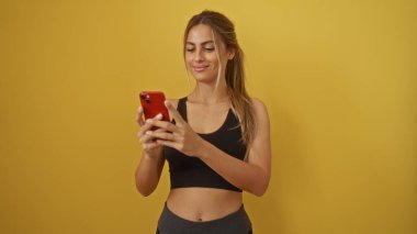 İzole edilmiş sarı arka planda akıllı telefon kullanan bir kadın, mesajlaşırken gülümseyen, sarı saçlı, sıradan kıyafetli, beyaz bir kadın..