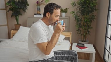 Orta yaşlı bir adam rahat bir yatak odasında kahve içiyor, yatakta oturuyor, günlük giysiler giyiyor, arka planda bitkiler ve pencereden süzülen güneş ışığı var..