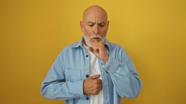 Orta yaşlı, sakallı, gri saçlı bir adam tek başına, parlak sarı bir arka planda dikilirken öksürüyor..