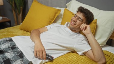 Genç bir İspanyol adam elinde uzaktan kumandayla yatakta dinleniyor. Sarı yastıklı ve ev dekorasyonlu rahat bir yatak odasında gülümsüyor..