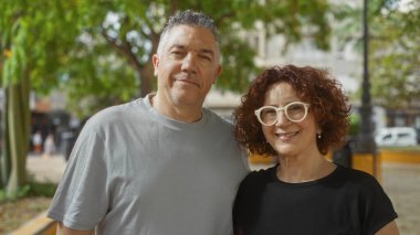 Arka planda yemyeşil bir yeşillik olan güneşli bir şehir parkında orta yaşlı bir çift birlikte gülümsüyor..