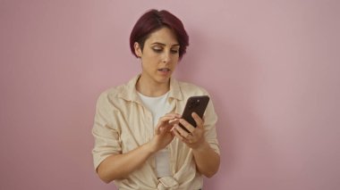 Kısa saçlı güzel beyaz bir kadın, izole edilmiş pembe bir arka planda dikiliyor, bej bir gömlek giyerek akıllı telefonuna dikkatle bakıyor..