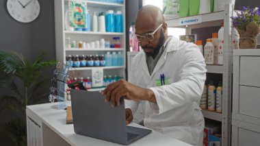 Eczanede dizüstü bilgisayarda çalışan genç Afrikalı bir adam. Etrafı çeşitli tıbbi ürünlerle çevrili beyaz bir ceket giyiyor.