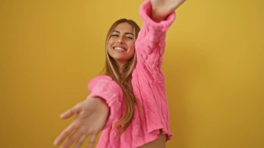 Neşeli bir şekilde gülümseyen, neşeli bir jestle elini uzatan, pembe giyimli, canlı sarı arka plan, mutluluk ve pozitifliği somutlaştıran bir kadın..