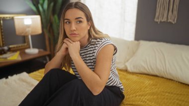 Genç sarışın bir kadın, rahat bir yatak odasındaki bir yatakta düşünceli bir şekilde oturur. İç hatları yumuşak ışıklandırma ve şık bir dekor içerir..