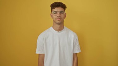 İzole edilmiş sarı bir arka planda duran gözlüklü genç İspanyol bir adam. Kendine güveni tam ve yakışıklı bir yüz ifadesi var..