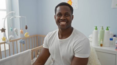 Kapalı alanda gülümseyen yakışıklı genç adam rahat bir yatak odasında bebek beşiğinin yanında oturuyor ve arka planda kişisel bakım malzemeleriyle sıcak ve davetkar bir atmosfer yaratıyor..