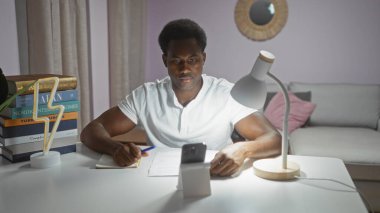 Oturma odasındaki bir masada kitap, lamba ve akıllı telefonuyla çalışan genç bir adam..