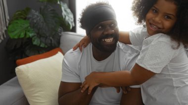 Baba ve kızı sıcak bir oturma odasında kucaklaşıyorlar. Sevgi ve samimiyet sergiliyorlar. Afro-Amerikan bir aile, kapalı alanlarda neşeli anlar yaratıyor..