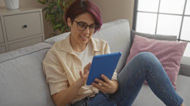 Kısa saçlı genç bir kadın modern oturma odasının içinde tablet kullanıyor..