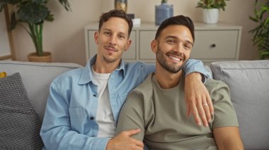 Sıcak bir oturma odasındaki kanepede oturmuş birbirine gülümseyen ve sarılan iki adam, rahat bir yuva ortamında sevgi ve aileyi somutlaştırıyor..