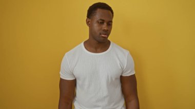 İzole edilmiş sarı arka planda poz veren, beyaz tişörtlü, düşünceli, çekici ve şık görünümlü genç Afro-Amerikalı bir adam.