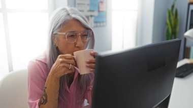 Ofisteki masasında oturan bir kadın, parlak bir iç mekanda bilgisayarla çalışırken kahve içiyor.