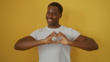Sarı duvara gülümseyen genç adam elleriyle kalp hareketi yapıyor, sıcaklığı ve mutluluğu minimalist bir ortamda taşıyor..