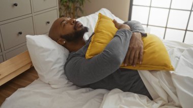 Genç, yakışıklı, Afrikalı, Amerikalı, kel, sakallı adam rahat bir yatak odasında bir yatakta yatıyor, sarı bir yastığa sarılıyor,.