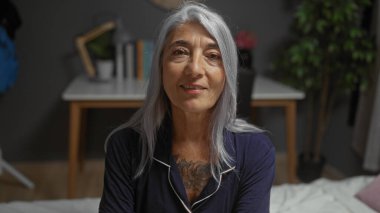 Güzel, gri saçlı bir kadın, rahat bir yatak odasında oturuyor, bir yatak odası ve iç mekan bitkileriyle, ev ortamında huzurlu bir gülümseme sergiliyor, sıcaklığını ve rahatlığını iletiyor..