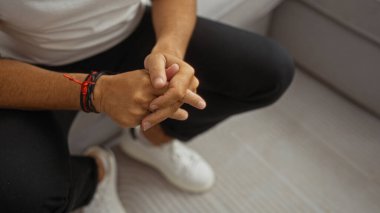 Oturma odasında elleri kucağında oturan adam bilezik takıyor ve beyaz ayakkabılı siyah pantolon giyiyor.