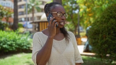 Güneşli bir günde arka planda binalar olan şehir parkında telefonla konuşan bir kadın.