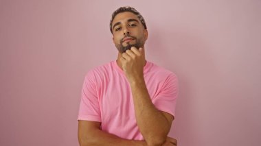 Sakallı ve pembe gömlekli genç bir Arap, pembe bir arka planda kollarını kavuşturarak düşünceli bir şekilde poz veriyor.