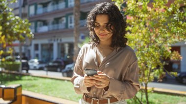 Genç, çekici bir İspanyol kadın şehir parkında telefonuyla mesajlaşırken gülümsüyor..