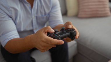 Oturma odasında oyun kontrolörüyle video oyunu oynayan genç bir adam, ev ortamında yakalanmış, ellerine odaklanmış ve gelişigüzel ayarlar yapıyor.