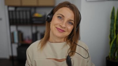 Kulaklık takan güzel sarışın bir kadın ofis ortamında çalışıyor. Arkasında bitki ve raflar var..