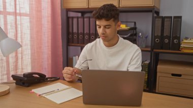 Ofis masasında çalışan Latin kökenli genç bir adam arka planda organize raflar olan bir dizüstü bilgisayar kullanıyor..