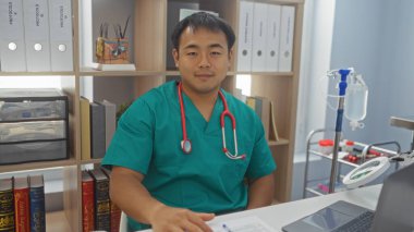 Genç, çekici, tıbbi önlüklü, boynunda steteskop olan Çinli bir adam klinikte iş yerinde oturuyor.