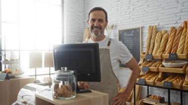 Orta yaşlı bir adam fırın tezgahında çalışıyor ve arka planda çeşitli ekmekler sergileniyor..