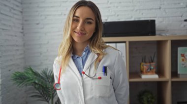 Modern bir klinikte gülümseyen, sarı saçlı ve boynunda steteskop olan çekici bir bayan doktor..