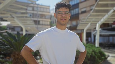 Gözlüklü yakışıklı, beyaz tişörtlü, kentin arka planında modern binalar olan bir parkta güvenle duran İspanyol bir adam..