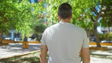 Genç bir adam yeşil ağaçlarla dolu bir parkta güneşli bir günde gölgenin altında beyaz bir tişört ve şehirde görünür bir kol dövmesi giyerek dikiliyor.