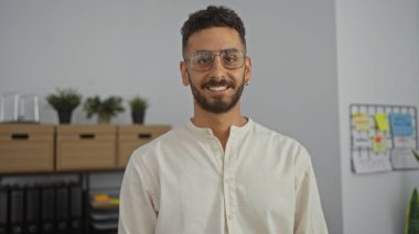 Ofis ortamında gülümseyen yakışıklı, gözlüklü ve sıradan giyinmiş, bitki ve ofis malzemelerinden oluşan yakışıklı bir İspanyol..