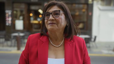 Kısa saçlı ve gözlüklü güzel, olgun bir İspanyol kadın şehir sokaklarında kırmızı ceket ve inci kolye giyerek dikiliyor..