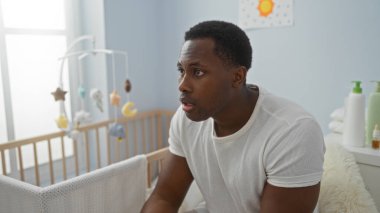 Bir adam beşikteki parlak bir yatak odasında oturuyor, nazik ve düşünceli görünüyor, yumuşak renkler ve bebek eşyalarıyla çevrili, huzurlu bir ev ortamını gösteriyor..