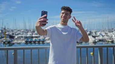 Genç İspanyol bir adam deniz kıyısındaki bir limanda selfie çekerken zafer jesti yapıyor..