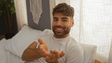 Sakallı, genç, İspanyol bir adam, gülümseyerek ellerini uzatıyor, rahat, aydınlık bir yatak odasında bir yatakta oturuyor..