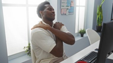 Young man embracing himself in an office, looking content, with a desk setup in front of him and plants on the windowsill clipart