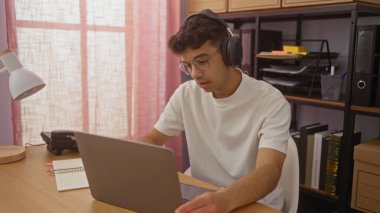 Kulaklık takan İspanyol bir genç ofis içinde dizüstü bilgisayarda çalışıyor. Rafları ve masa lambası var..