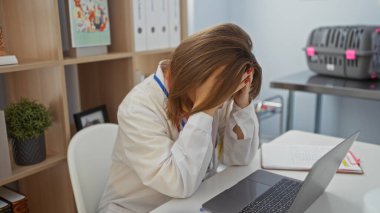 Klinik odasında hayal kırıklığına uğramış sarışın bir kadın veteriner, elinde bir dizüstü bilgisayarla, etrafında ofis dosyaları ve veteriner aletleriyle otururken kafasını elinde tutuyor..