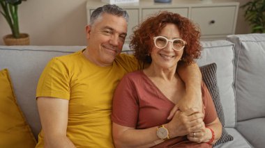 Middle-aged couple sitting on a couch in a cozy living room, smiling and embracing each other, showing love and happiness indoors. clipart