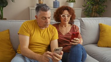 Oturma odalarında birlikte oturan orta yaşlı bir çift, her ikisi de akıllı telefonlarına odaklanmış, sarı yastıklı rahat bir kanepede dinleniyorlardı.