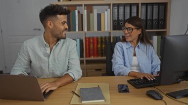 Kadın ve erkek bir ofiste birlikte çalışıyor, sıralarında dizüstü bilgisayarları ve kitaplıklarıyla gülüyor ve etkileşime geçiyorlar, profesyonel ve işbirlikçi bir ortam sergiliyorlar.