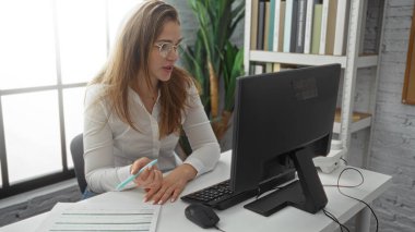Ofisteki bir masada çalışan bir kadın, bilgisayar ve evrak işleriyle uğraşıyor, kitaplıklar ve doğal ışıklandırmalarla profesyonel bir iç mekan sergiliyor..