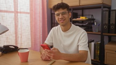 Ofisteki genç İspanyol bir adam elinde akıllı bir telefon, gülümsüyor ve modern bir iş ortamı yaratıyor..