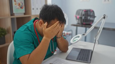 Yeşil üniformalı genç Asyalı bir adam, veteriner kliniğinde çalışırken, dizüstü bilgisayarı ve masada belgelerle stresli görünüyor..